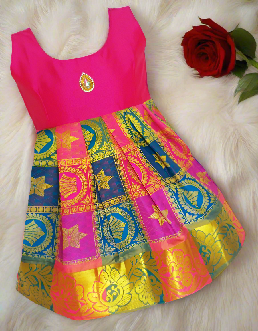 A silk frock for girls with a blue, pink, and yellow geometric pattern and golden zari weaving, displayed on a white furry surface with a red rose beside it.